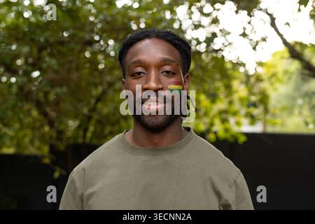 African American man posing in garden in olive crewneck, green-yellow cheek paint, nose-ring Stock Photo