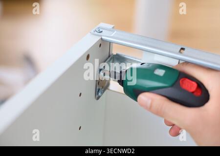 A woman using a compact cordless electric screwdriver to assemble white wooden furniture, focusing on a metal corner bracket. Close up. Stock Photo