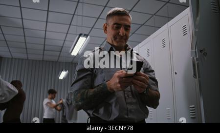 Male Security Officer With Arm Tattoos Hangs ID Badge on Locker Door, Takes Out Smartphone, and Checks For Missed Calls and Messages After Finishing Workday. Shift End Concept in Staff Dressing Room. Stock Photo