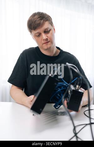 Young man in black shirt sitting at white table concentrating on tangled wires. He holding electronic devices, attempting to solve technical problem Stock Photo