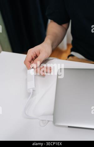 Close-up of male hand plugging white internet adapter into modern laptop. Minimalist workspace with focus on technology, connection, remote work Stock Photo