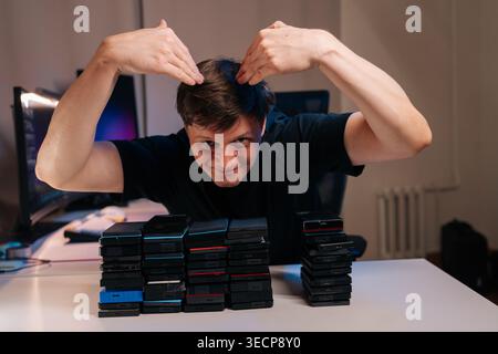 Man celebrating significant amount of digital data storage, posing playfully behind two tall stacks of portable hard drives on desk in home office Stock Photo