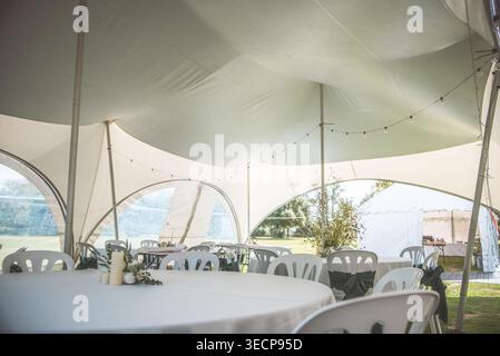 A beautiful outdoor wedding reception is set up under a large white tent with string lights and round tables Stock Photo