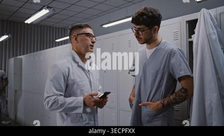 After Busy Workday, Two Male Doctors Relax by Grey Metal Lockers, Viewing Sport News on Smartphone and Chatting About Personal Interests in Locker Room. Concept of Shift End and Employee Wellness. Stock Photo