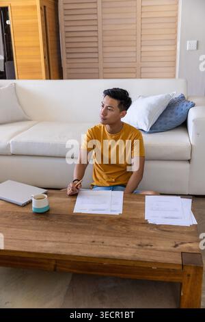 Adult Asian man sitting on floor in living room wearing mustard T-shirt, reviewing papers with pen Stock Photo