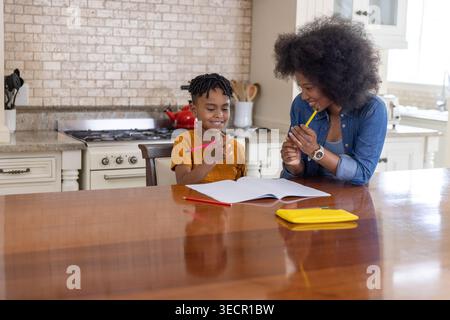African American mother and son leaning over notebook at kitchen table using colored pencils Stock Photo