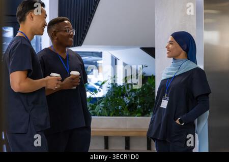 Diverse medical coworkers standing and talking in clinic lobby wearing navy scrubs holding coffee Stock Photo