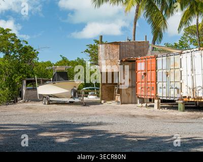 USA, Islamorada, FL - April 18, 2026: A white motorboat on a trailer stands on a gravel lot near several shipping containers and tropical palm trees. Stock Photo