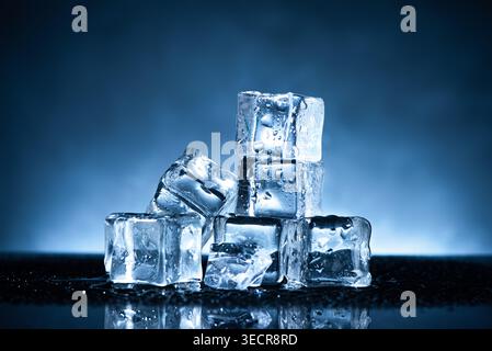 Ice cubes melting, showing water droplets on a reflective dark surface against a blue background Stock Photo
