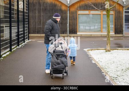 A parent pushes a stroller while a small toddler in a blue winter jacket walks ahead on a wet pathway during winter. The scene shows everyday family l Stock Photo