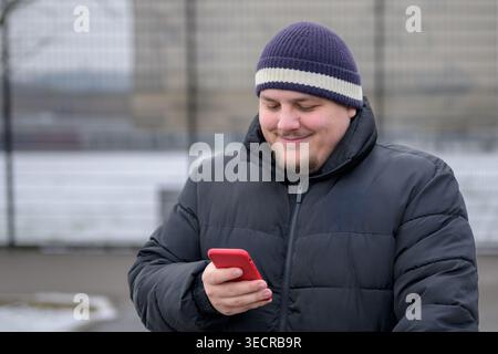 Smiling young man wearing a winter jacket and knit beanie while looking at his smartphone outdoors in an urban winter setting. Stock Photo