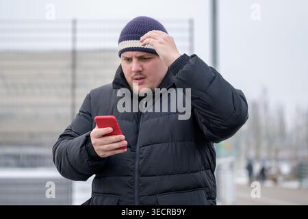 Young man wearing a black winter jacket and knit hat looking at his smartphone outdoors on a cold winter day. Stock Photo