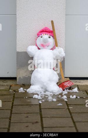 Cute handmade snowman standing outdoors against a wall, holding a broom and wearing a pink knitted hat on a winter day. Stock Photo