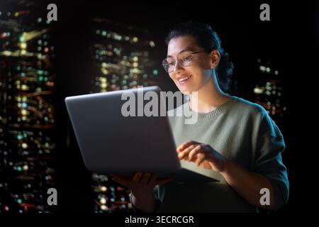 A confident young woman IT professional in glasses stands in a high-tech server room, smiling while working on her laptop. The dim, atmospheric lighti Stock Photo