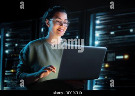 A young woman in glasses works on a laptop in a high-tech server room, illuminated by the glow of server racks. Her focused and positive expression co Stock Photo