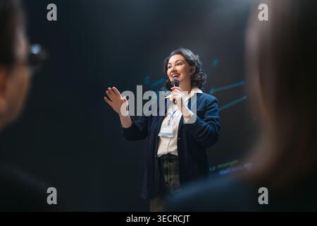 Dynamic image of a confident female speaker engaging an audience during a professional business conference. The atmosphere radiates inspiration and em Stock Photo