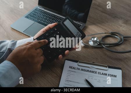 Hand using calculator on medical insurance claim form beside laptop and stethoscope performing financial calculation for healthcare expense budgeting, Stock Photo