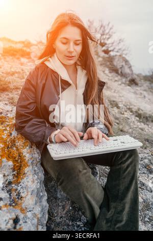 Woman keyboard outdoors sitting on rocky terrain at sunset using wireless technology in nature during scenic golden hour Stock Photo