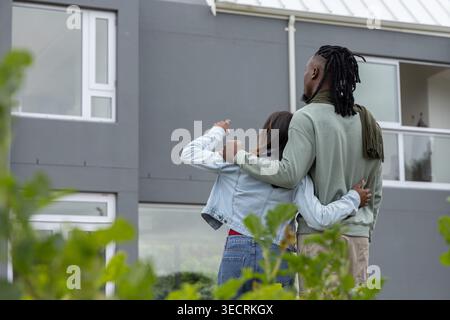 Diverse couple standing outside modern grey building pointing at balcony wearing denim jacket scarf Stock Photo