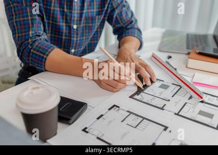 A young male architect is working with blueprints at his desk, using rulers and drawing tools to refine details. He appears focused and engaged, balan Stock Photo