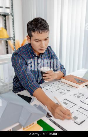 A young architect is working at his desk, reviewing blueprints, using a calculator, and checking a house model. He focuses on precision and detail whi Stock Photo