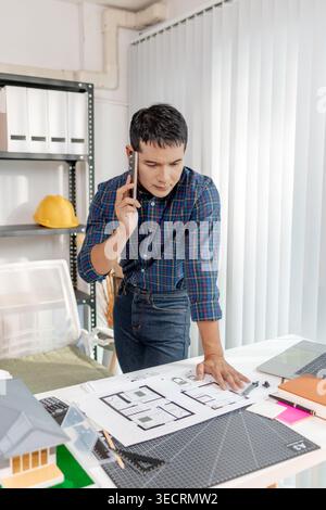 A young architect is standing at his desk while discussing a project on the phone, carefully reviewing construction plans with focus and attention in Stock Photo