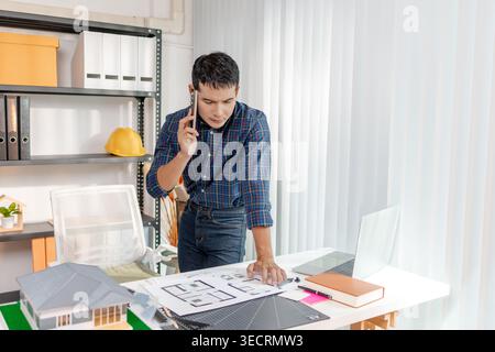 A young architect is standing at his desk while discussing a project on the phone, carefully reviewing construction plans with focus and attention in Stock Photo