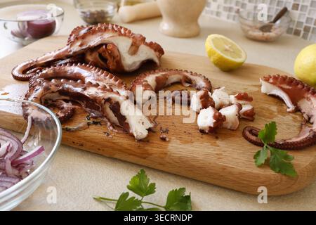 Freshly cooked octopus sliced on wooden board in kitchen. Chef prepares ingredients for healthy mediterranean seafood salad with lemon and red onion Stock Photo