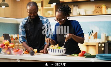 Happy black man squeezing lemon juice over fresh vegetables, preparing a tasty salad together with girlfriend. Seasoning lunch meal with bio organic ingredients, relaxed home moment. Stock Photo