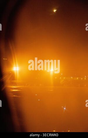 Nighttime view from an airplane window shows a foggy airport apron with glowing runway lights and reflections on wet pavement, creating a warm, hazy t Stock Photo