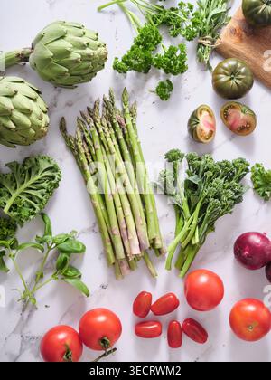 Various fresh vegetables are arranged on a marble kitchen countertop. This colorful display shows asparagus, tomatoes, and herbs, suggesting preparati Stock Photo