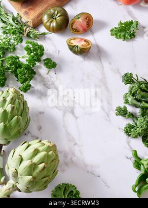 Various vegetables are arranged on a marble kitchen countertop. Fresh greens and sliced tomatoes highlight the healthy ingredients ready for cooking. Stock Photo