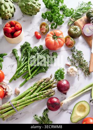 Various vegetables are placed on a marble kitchen countertop. They include tomatoes, onions, asparagus, and herbs, ready for preparation for a healthy Stock Photo