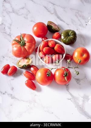 Top view of assorted fresh tomatoes including cherry, grape, heirloom and green varieties, arranged around a bowl on a white marble background for coo Stock Photo