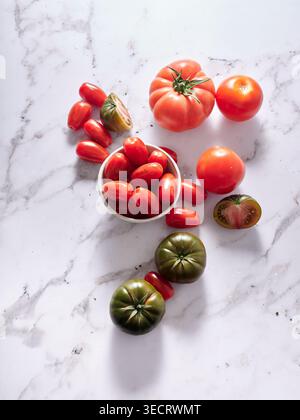 Top view of fresh assorted tomatoes including heirloom, vine and cherry varieties in a bowl on white marble surface, vibrant red and green produce. Stock Photo