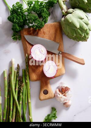 Various vegetables are placed on a marble kitchen countertop. A knife rests on a wooden cutting board next to chopped onions, asparagus, and garlic, s Stock Photo