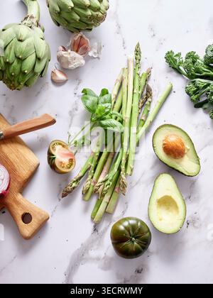 Various vegetables are laid out on a marble countertop. Ingredients include artichokes, asparagus, avocados, and herbs for cooking in the kitchen. Stock Photo