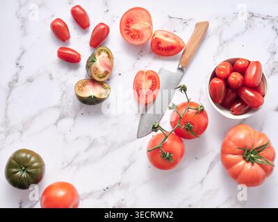 Various vegetables are set on a marble countertop in a kitchen. Sliced tomatoes, whole tomatoes, and a knife are visible. Natural light fills the area Stock Photo