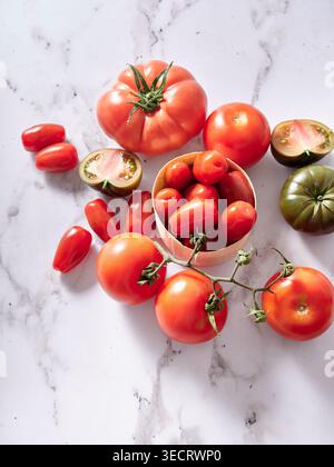 Fresh heirloom and cherry tomatoes arranged on a white marble surface, featuring vine tomatoes, sliced green heirloom halves, and a cup of red grape t Stock Photo