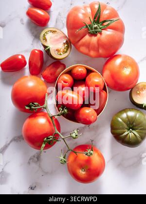 Fresh heirloom and cherry tomatoes arranged on a white marble surface, featuring vine tomatoes, sliced green heirloom halves, and a cup of red grape t Stock Photo