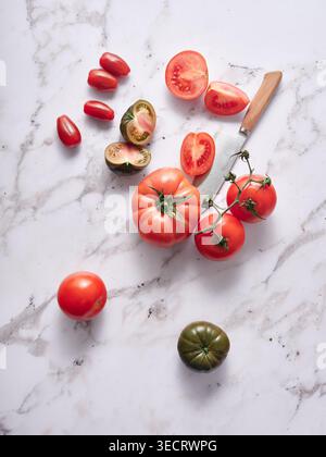 Assorted fresh tomatoes, including heirloom, vine and cherry varieties, arranged with sliced halves and a knife on a white marble surface, minimal foo Stock Photo