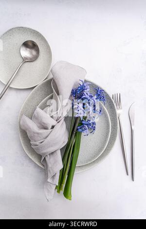Minimal table setting featuring stacked ceramic plates, silver cutlery, and a soft linen napkin, accented by a fresh blue hyacinth on a light backgrou Stock Photo