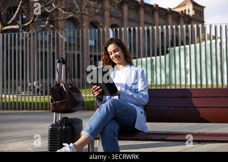 Smiling business traveler sits on an outdoor bench using a tablet beside a suitcase and handbag, working remotely while waiting in a city setting duri Stock Photo