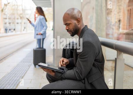 Focused businessman in suit using a tablet at a transit stop while colleague with suitcase checks phone in background, capturing business mobility and Stock Photo