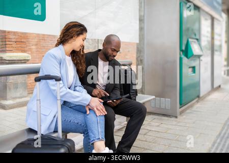 Business travelers couple sitting at a station bench with luggage, checking a tablet together while waiting for transport, showcasing modern mobility Stock Photo