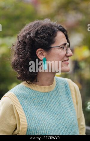 A mature woman with curly hair and stylish earrings enjoys a pleasant day in an urban environment. She appears content and relaxed, embodying a vibran Stock Photo