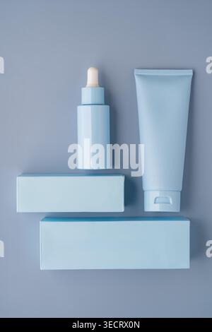 Minimal flat lay of pastel blue cosmetic packaging featuring a serum dropper bottle, cream tube, and blank boxes on gray background, ideal for skincar Stock Photo