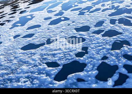Closeup of soap foam and water droplets spread across car glass during washing, creating an abstract wet texture with blue reflections and glossy shin Stock Photo