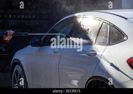 Close-up of a person using a pressure washer to clean a white car, water spray and mist catching sunlight on the side window and door, concept for det Stock Photo