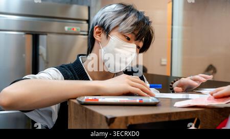 A focused young worker in a Taiwanese restaurant, wearing a mask, handles customer orders at the counter, showcasing attention and dedication in a mod Stock Photo